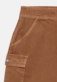 Brune bomullsbukser med en frontlomme og en sidetaskepocket med trykknapp-lukking. Stoffet har en teksturert overflate.