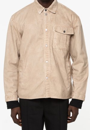 Hombre con chaqueta beige abotonada con bolsillo en el pecho, camisa negra de manga larga debajo y pantalones negros sobre fondo blanco.