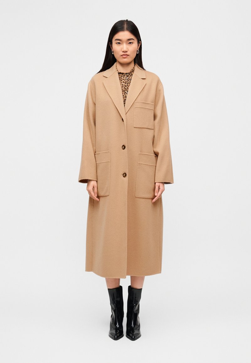 Μπεζ μάλλινο παλτό με μακριά, ίσια σχεδίαση, δύο μπροστινές τσέπες και κλείσιμο με ένα κουμπί, σε συνδυασμό με μαύρες μποτάκια μέχρι τον αστράγαλο.