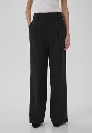 Fekete magas derekú, bő szárú nadrágot és fehér ujjatlan felsőt viselő személy, zsebébe dugott kézzel áll, egyszerű háttér előtt.