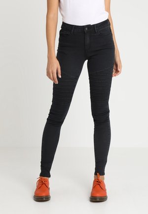 Femme portant un jean skinny côtelé noir, un haut blanc et des chaussures à lacets orange vif, debout sur un fond clair uni.