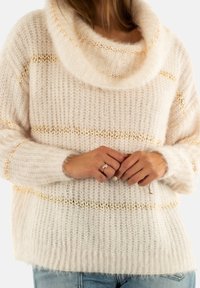 Frau, die einen weichen, cremefarbenen Strickpullover mit goldenen horizontalen Streifen trägt, steht mit gefalteten Händen vor sich und trägt einen Ring.