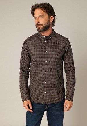 Chemise marron à manches longues avec un petit motif géométrique, boutons blancs, associée à un jean bleu. Texture de tissu lisse.