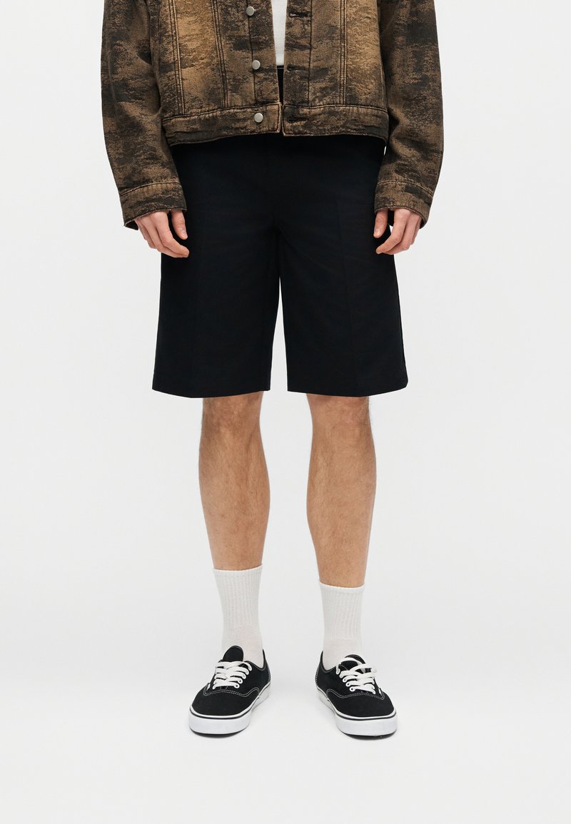 Persona che indossa pantaloncini neri fino al ginocchio, calzini bianchi corti, scarpe da ginnastica nere e una giacca marrone testurizzata con bottoni d'argento.