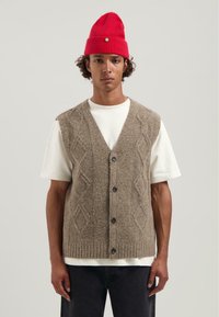 Junger Mann mit einer roten Strickmütze, einem weißen kurzärmligen Hemd und einer grauen, geknöpften Wollweste mit Rautenmuster.