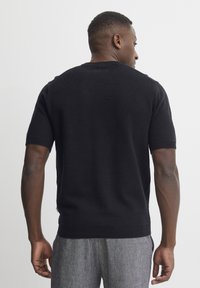 Pulover scurt negru cu mânecă scurtă, gât rotund, textură tricotată și tiv ondulat. Purtați cu pantaloni texturați de un gri deschis.