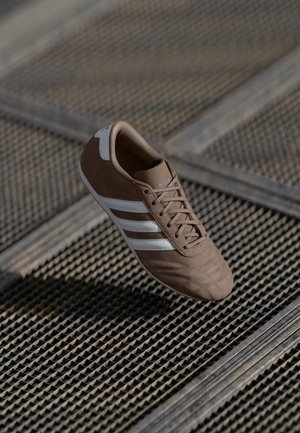 Bruine sneaker met witte strepen die zweeft boven een getextureerd metalen oppervlak in natuurlijk licht.