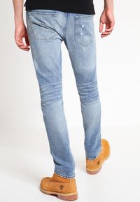 Persona vistiendo unos jeans destruidos de color azul claro y botas Timberland color beige, vista desde atrás sobre un fondo blanco.