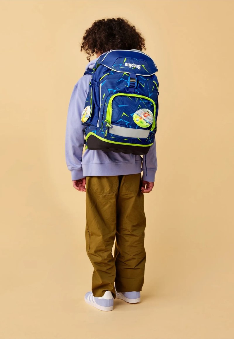 Blauer und grüner gemusterter Rucksack mit zwei runden Patches, verstellbaren Trägern und einer Vordertasche. Von einem Kind in helllila und khakifarben getragen.