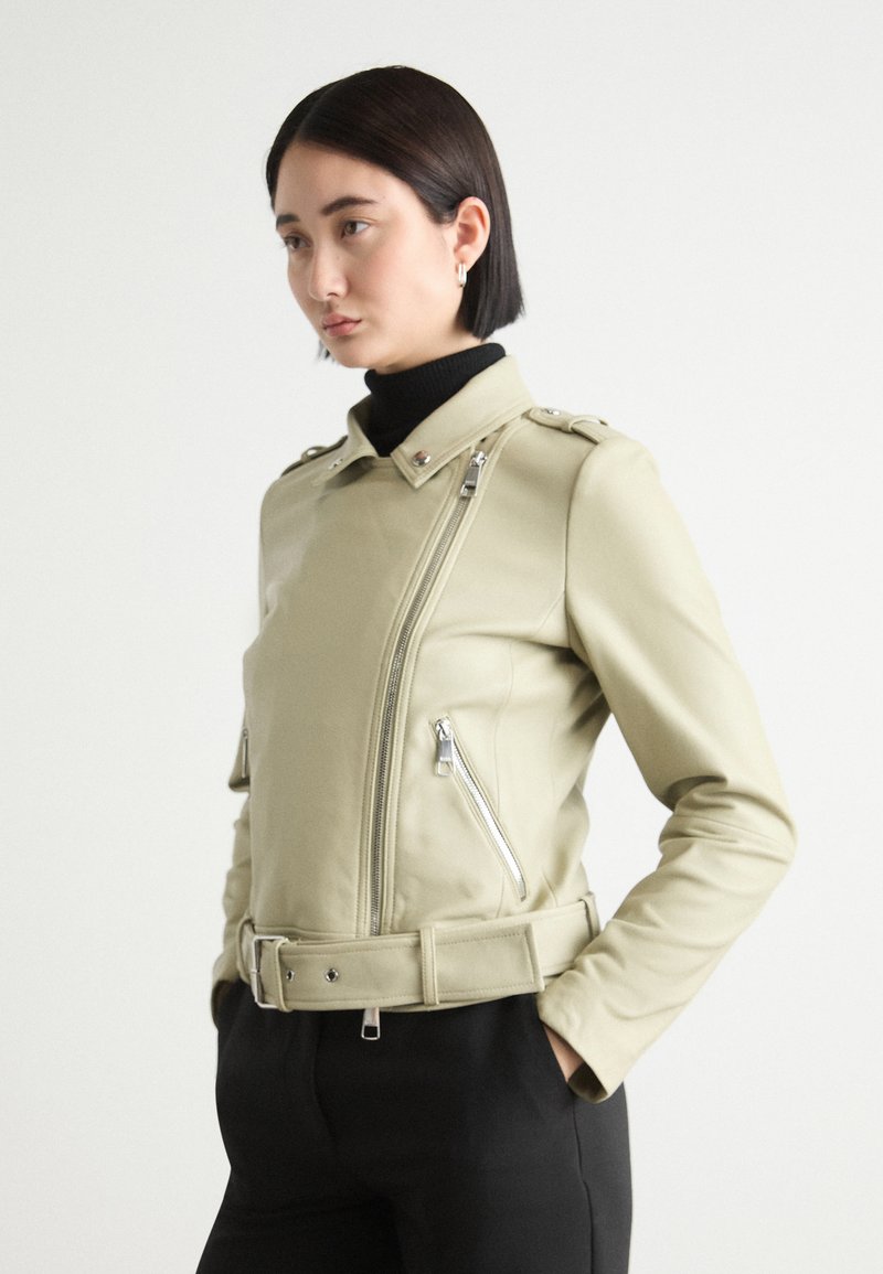 Jachetă din piele ecologică de culoare verde deschis, ajustată, cu fermoar asimetric, guler cu capse și talie cu curea. Prezintă epoleti pe umeri și fermoare laterale.
