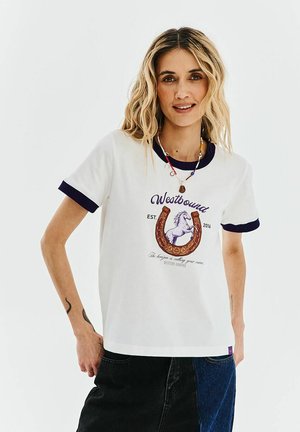 Kobieta o falowanych blond włosach, ubrana w biały T-shirt z granatowymi wykończeniami i grafiką konia z podkową, w połączeniu z czarno-niebieską spódnicą patchworkową.