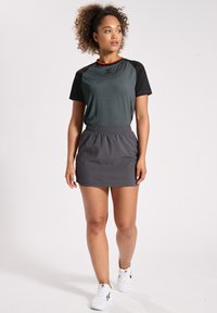 Grå träningskjol kombinerad med en kortärmad mörkgrön och svart topp, detaljerad med röda kantband. Modellen bär vita sneakers.