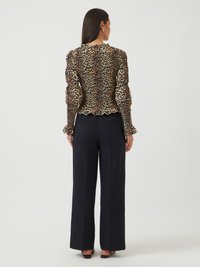Blouse en imprimé léopard avec manches bouffantes, tissu texturé et ourlet à volants, associée à un pantalon noir à jambe large. Vue de dos, cheveux lisses et boucles d'oreilles visibles.