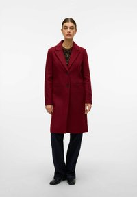 Cappotto di lana bordeaux con colletto a revers, chiusura con un bottone e due tasche frontali. Abbinato a pantaloni larghi scuri.