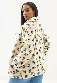 Kukallinen fleece-takki vaalean kermanvärisenä, jossa on monivärisiä kukkakuoseja. Varustettu kauluksella ja sivutaskuilla. Pehmeä, tekstuurinen kangas.