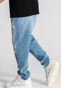 Jeans de mezclilla azul claro con un corte relajado, que presentan detalles desgastados y puños ajustados, llevados con zapatillas blancas que muestran una gran marca.