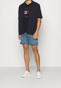 Camiseta oscura de manga corta sobre una camiseta gráfica negra, combinada con pantalones cortos de mezclilla y zapatillas blancas. Diseño simple, ajuste casual.