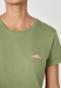 Detailaufnahme einer Person in einem grünen T-Shirt mit einem kleinen, bestickten pinken Leoparden und dem Text "out for SUMMER" auf der linken Brust.