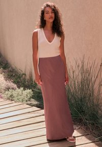 V-hals kjole med tekstureret cremefarvet top og flydende mauve bund. Lang, slank silhuet; kombineret med hvide sandaler med stropper. Naturlig baggrund.