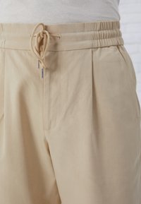 Pantalon beige avec taille élastique et fermeture à cordon. Présente deux plis avant et un tissu lisse et léger.