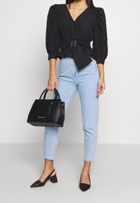 Blusa negra ajustada con mangas abullonadas, jeans de tiro alto en azul claro y un bolso de cuero negro. Tacones de punta negra con talón abierto.