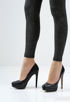 Pompes à talons hauts en cuir noir avec un bout arrondi et une semelle à plateforme, assorties à des leggings noirs texturés. Finition lisse et design épuré.