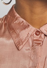Primo piano di un collo di una persona che indossa una camicia color rosa luccicante con il colletto e il primo bottone allacciato, con un orecchino a cerchio di argento visibile.