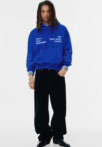 Blauwe hoodie met witte tekst, voorzien van een kangoeroezak en grote manchetten. Gecombineerd met brede zwarte ribfluwelen broek en zwarte laarzen.