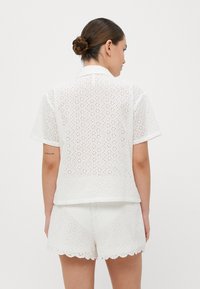Hvitt bomullsdress i to deler med kortermet topp og shorts med bølgeskjørt, begge med intrikate hulmønster.