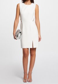 Robe blanche sans manches avec une coupe droite, des fentes latérales et des accents scintillants. Associée à un sac à main métallique argenté et des talons.