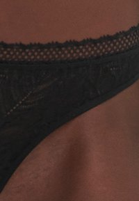 String en dentelle noire avec un motif floral délicat, doté d'une ceinture élastique festonnée et d'une texture transparente. Mettez l'accent sur les détails intriqués.