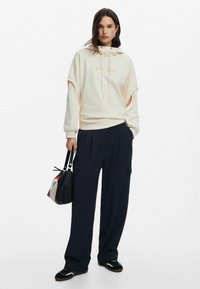 Cream hoodie with an embroidered logo, wide-cut sleeves, paired with dark navy cargo pants and black shoes. Carrying a multi-colored bag.