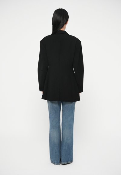 Černé sako s vycpávkami na ramenou, strukturovaným střihem a dlouhými rukávy. Spárováno se světle modrými rozšířenými džínami, vytváří výraznou siluetu.