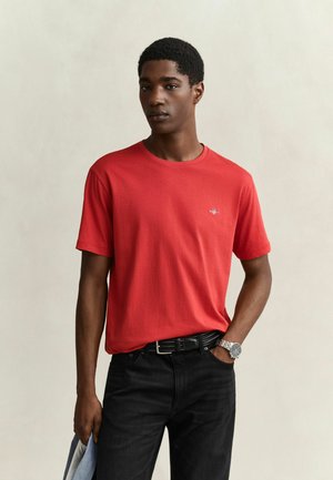Jonge man in een rood T-shirt, zwarte broek, zwarte riem en een zilveren horloge, staand met één hand in zijn zak en met de andere hand kleding vasthoudend.