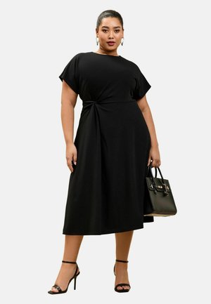 Žena stojaca v čiernych midi šatách so stranovým uzlom, čiernych sandáloch na vysokom podpätku, zlatých kruhových náušniciach, držiaca čiernu kabelku na bielom pozadí.