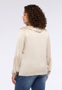 Beżowa jedwabna bluzka z długimi rękawami i marszczonym kołnierzem, o gładkiej fakturze i lekko luźnym kroju, zestawiona z niebieskimi dżinsami.