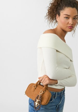 Borsa a tracolla in suede beige con doppi manici e pendente. Il modello indossa un top bianco a spalla scoperta con righe e jeans in denim blu.