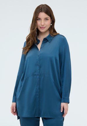 Frau mit langen braunen Haaren, die eine locker sitzende satinartige, tealfarbene Bluse mit Knöpfen und passende Hosen trägt, vor einem schlichten Hintergrund stehend.