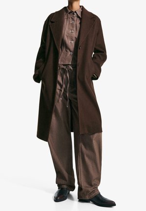 Personne portant un long manteau marron foncé sur une chemise marron boutonnée et un pantalon marron ample avec un cordon de réglage, associé à des chaussures noires.