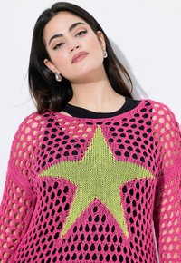 Rosa Häkelpullover mit einem großen gelben Sternmuster, ausgestattet mit offenem Strick und langen Ärmeln, über einem schwarzen Oberteil getragen.