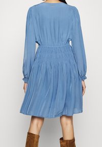 Kvinna som bär en blå långärmad klänning med samlad midja och manschetter, i kombination med bruna mocka knälånga stövlar, sett bakifrån.