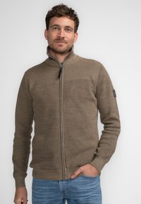 Suéter con textura en tono marrón y cremallera, con un cuello alto y puños acanalados, que presenta un diseño sutil en la parte frontal y un parche con el logo en la manga.