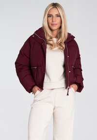 Bordowa kurtka puchowa z kapturem, z metalowymi suwakami i kieszeniami. Nosi się ją na jasnym topie i jasnych spodniach, ukazując luźny krój.