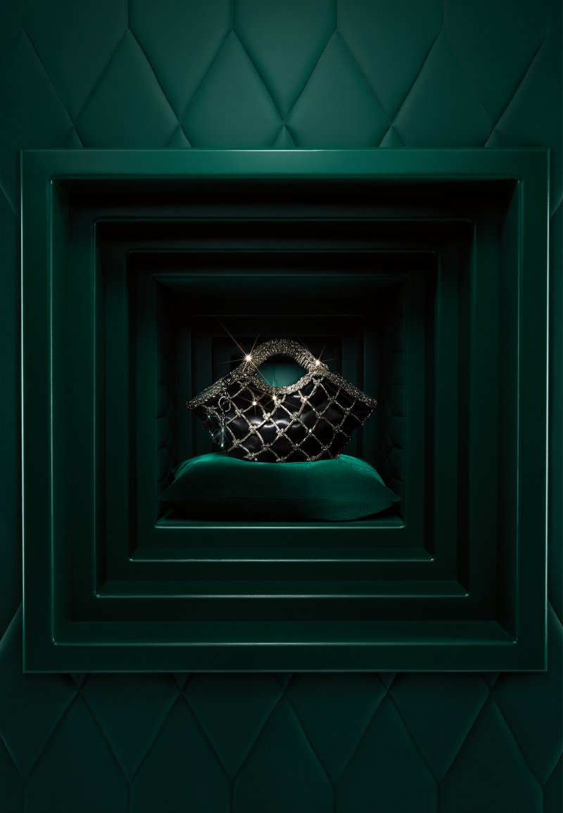 Schwarze gesteppte Handtasche mit metallischen Akzenten, präsentiert auf einem grünen Samtkissen, umrahmt von geschichteten grünen geometrischen Hintergründen.