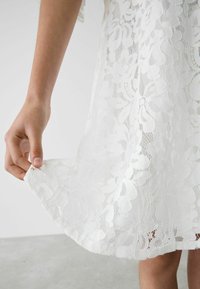 Witte kanten jurk met bloemmotieven, transparante textuur en een scalloped zoom. Hand houdt de stof aan de onderkant vast voor detailaccentuering.