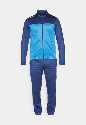 Tuta sportiva con una combinazione di colori blu e blu scuro, giacca con zip e colletto alto, polsini elasticizzati e pantaloni tapered abbinati con tasche laterali.