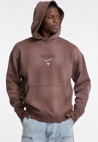 Felpa marrone con cappuccio, caratterizzata da una tasca a marsupio frontale, polsini a costine e logo "GUESS originals" con una grafica triangolare sul petto.