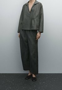 Tenue en tissu gris texturé comprenant une veste ample et courte avec un col en V et un détail noué, associée  à un pantalon à jambes larges et des chaussures foncées.