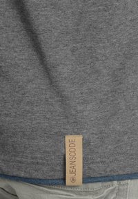 Graues T-Shirt aus einem weichen, strukturierten Stoff mit einem sichtbaren "JEAN SCODE" Markenetikett an der unteren Seite, unterstrichen von einem blauen Akzent.
