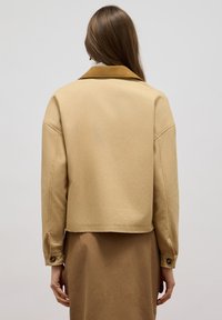 Beige cropped jasje met een bruine kraag, knoopsluiting bij de mouwen en een glad, mat oppervlak. Heeft een eenvoudig, minimalistisch ontwerp.